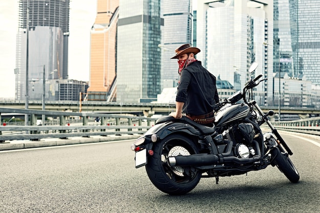 Ritratto di uomo elegante vestito con una giacca di pelle nera e bandana seduto sulla sua moto retrò su misura, guardando lontano, edifici della grande città sullo sfondo.