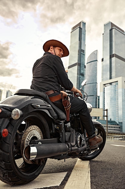 Portrait of stylish male dressed in a black leather jacket and bandana with gun sitting on his custommade retro motorcycle looking away buildings of the big city on background