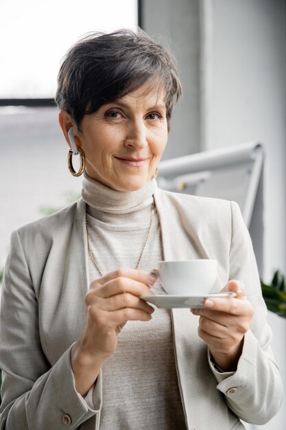 Портрет стильной зрелой деловой женщины в офисе с наушниками, кофейной чашкой и улыбкой в камеру.