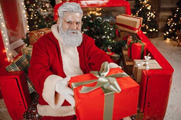 Photo portrait of a stylish santa claus with a gift in his hands, a lot of gifts in the back of a car. preparing gifts for the celebration of christmas and new year