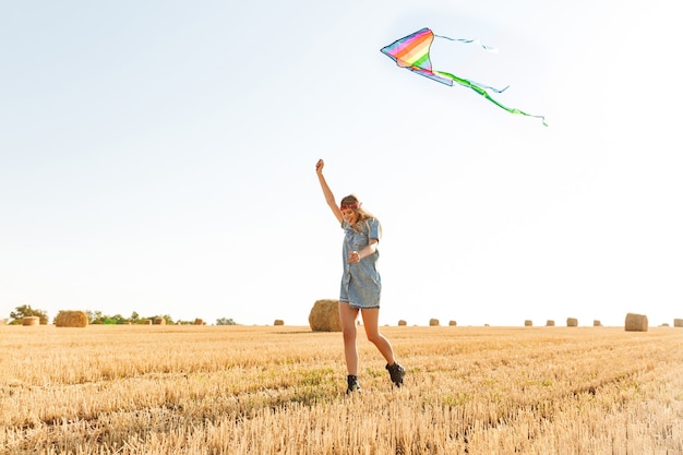 Portrait of stylish woman 20s smiling and playing with flying kite during walk through golden field, during sunny day