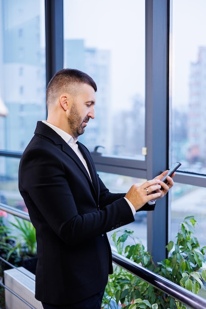 Portrait of stylish young businessman standing in large modern office high on top floor and looking at phone