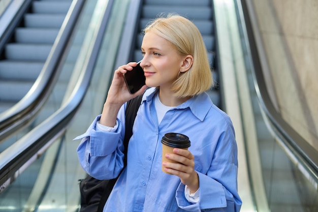 Photo portrait of stylish young woman with cup of coffee standing near escalator in city centre answer