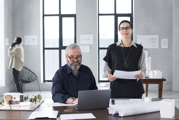 Portrait of successful architects looking at camera while working with blueprints and computer at the table at office