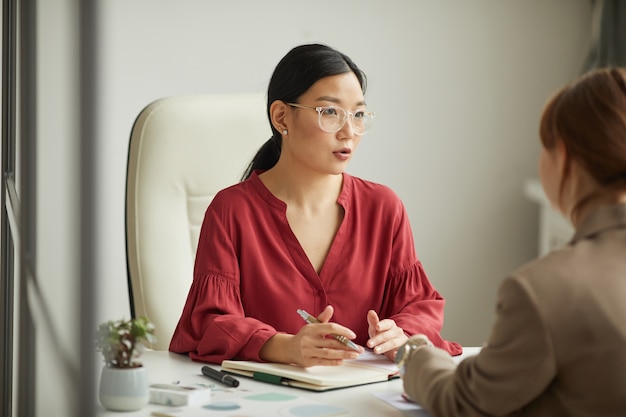 Portrait of successful Asian businesswoman talking to client while working at desk in white office cubicle, copy space