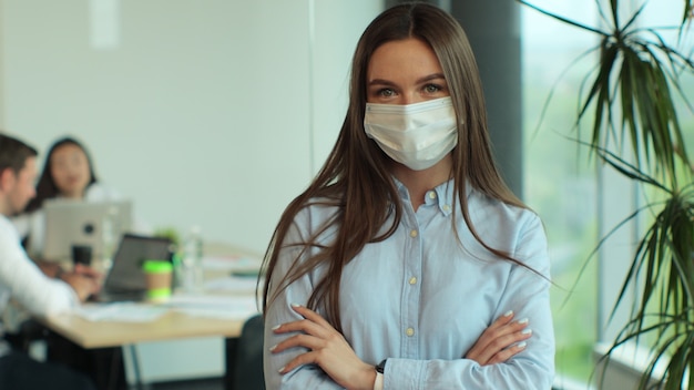 現代の会議室に立っている現代のオフィスの職場の若い女性で交差するカメラの腕を見て医療マスクで成功した美しいエグゼクティブ実業家スマートカジュアルウェアの肖像画