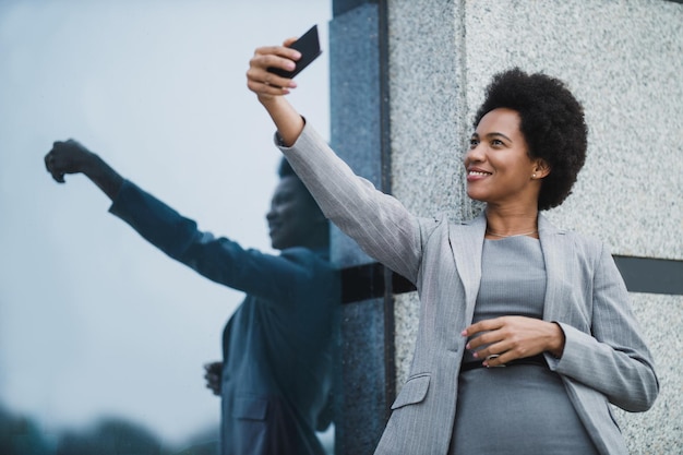 Portrait of a successful black business woman taking photos
with her smartphone during quick break in front a corporate
building.