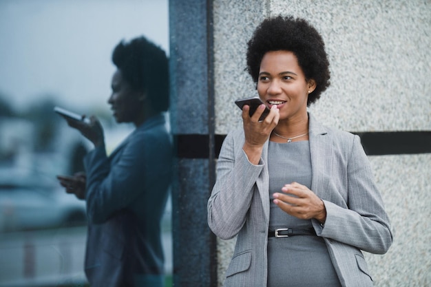 Portrait of a successful black business woman talking voice message on a smart phone during quick break in front a corporate building.