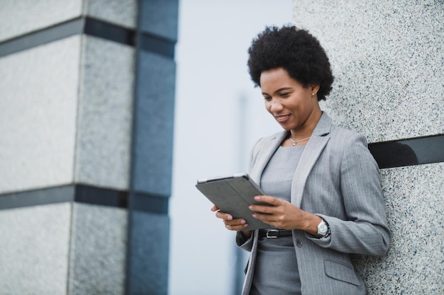 Портрет успешной чернокожей деловой женщины, использующей приложение на цифровом планшете во время быстрого перерыва перед корпоративным зданием.