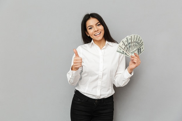 Portrait of successful brunette woman wearing formal outfit holding lots of dollars in hand and showing thumb up, isolated over gray wall