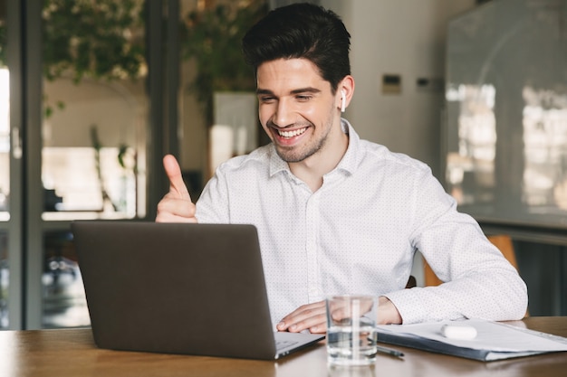 Portrait of successful businessman 30s wearing white shirt laughing and showing thumb up at laptop in office, during video conference or call using bluetooth earbud