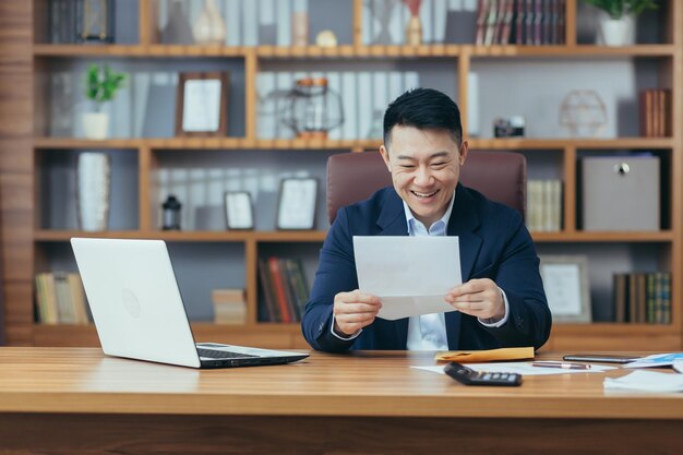 Ritratto di un uomo d'affari di successo asiatico ha ricevuto una lettera di approvazione dalla banca per estendere la linea di credito un uomo felice che lavora al tavolo con in mano una busta