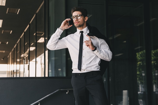 Портрет успешного бизнесмена, одетого в строгий костюм, стоящего у стеклянного здания с курткой через плечо и разговаривающего по мобильному телефону