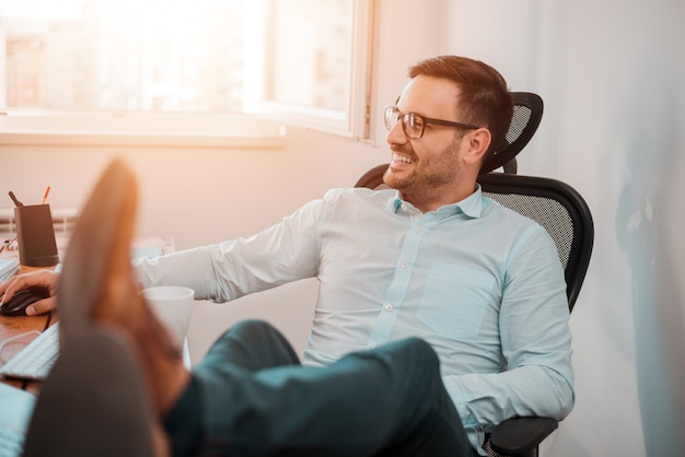 Portrait of successful businessman having rest on workplace in office.