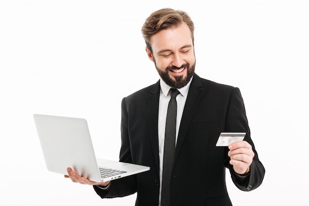 Portrait of successful businessman smiling and holding laptop and plastic credit card in hands, isolated over white wall