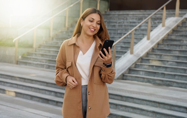 Portrait of successful businesswoman holding mobile phone while walking outdoors near financial office young woman entrepreneur using smartphone