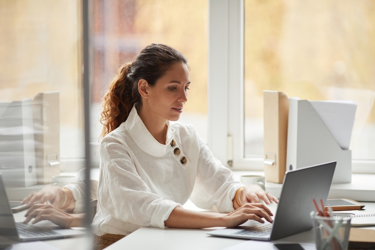 Photo portrait of successful businesswoman using laptop while sitting at desk by window and enjoying work in office, copy space
