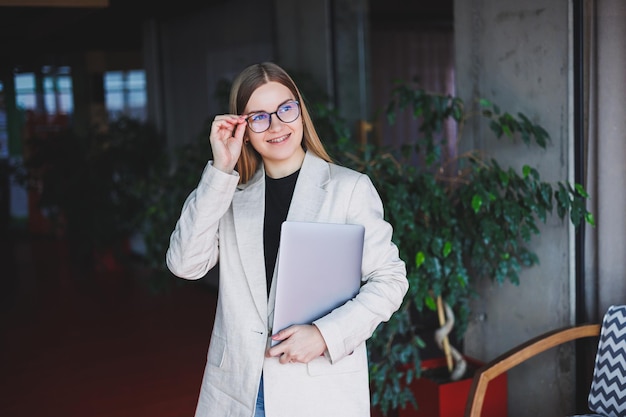 Portrait of a successful manager in the office smiling in during the working day happy woman with laptop enjoying time in modern office Blondhaired woman in a beige jacket and glasses