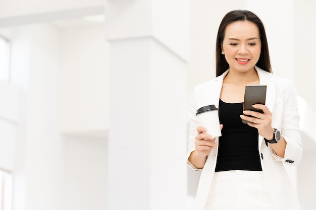 Портрет успешной азиатской бизнес-леди средних лет, держащей смартфон и кофе, улыбаясь в камеру