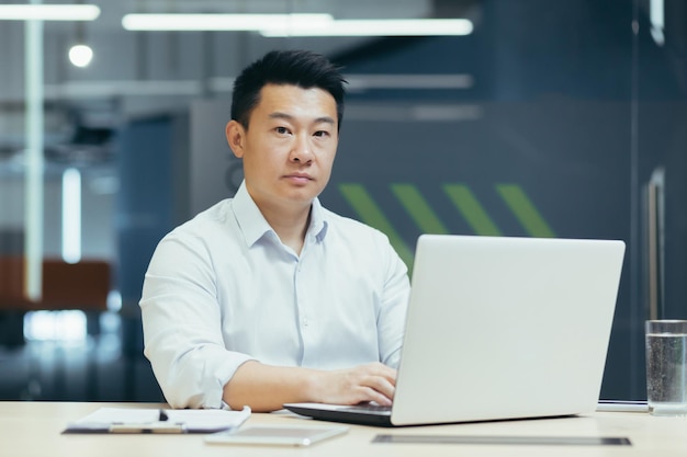 Portrait of successful and serious asian businessman man working in modern office typing on laptop