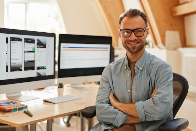 Portrait of successful software developer in eyeglasses standing with arms crossed and smiling at camera at office