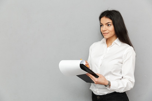 Portrait of successful woman with long dark hair wearing businesslike outfit holding clipboard with papers and writing down notes in documents, isolated over gray background