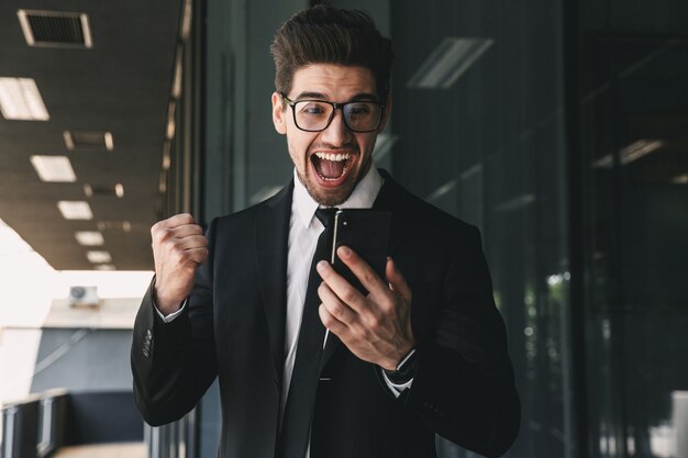portrait of successful young businessman dressed in formal suit standing outside glass building, and holding mobile phone