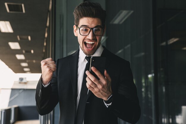 Portrait of successful young businessman dressed in formal suit standing outside glass building, and holding mobile phone