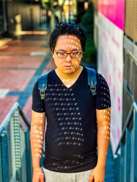 Photo portrait of sun dappled young asian man in eyeglasses standing on staircase in the city