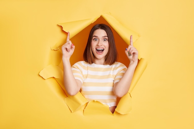 Portrait of surprised amazed woman stands in torn paper hole pointing fingers up presenting copyspace looking through breakthrough of yellow background