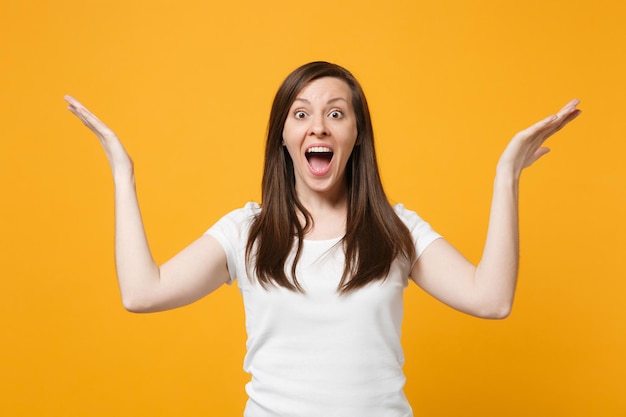 Portrait of surprised amazed young woman in casual clothes keeping mouth open, spreading, pointing hands aside isolated on yellow orange wall background. People lifestyle concept. Mock up copy space.