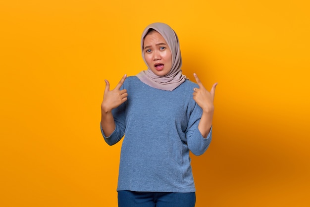 Portrait of surprised Asian woman standing and pointing finger herself over yellow background