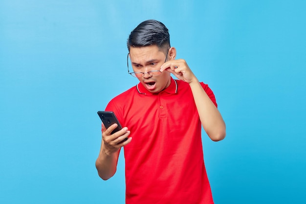 Портрет удивленного азиатского молодого человека, держащего очки и читающего входящее сообщение на смартфоне на синем фоне