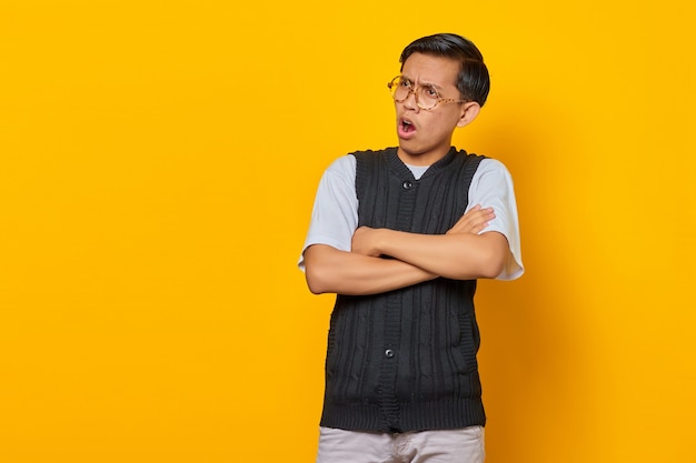 Portrait of surprised asian young man with crossed arms isolated on yellow background