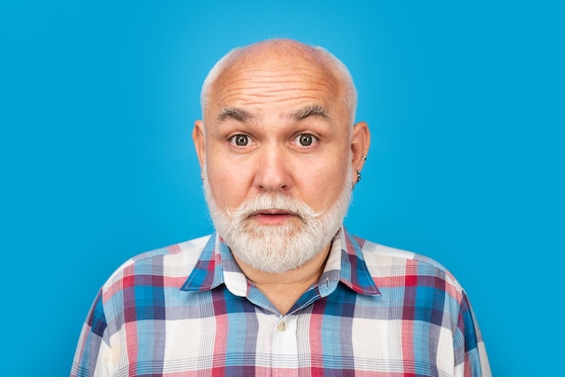 Portrait of an surprised bald old mature senior man with grey beard in shirt isolated on blue studio