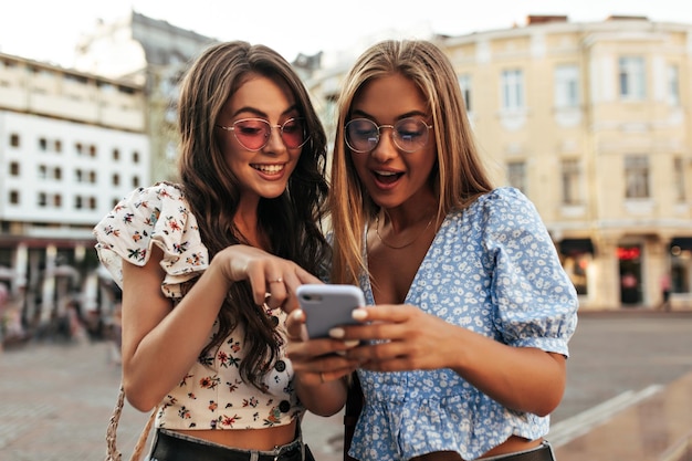 Ritratto di donne brune e bionde sorprese che leggono messaggi sul cellulare fidanzate in eleganti camicette estive e occhiali da sole colorati parlano all'esterno