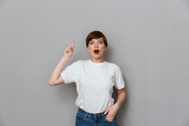 Portrait of surprised brunette woman wearing casual t-shirt pointing finger upward at copyspace isolated over gray wall