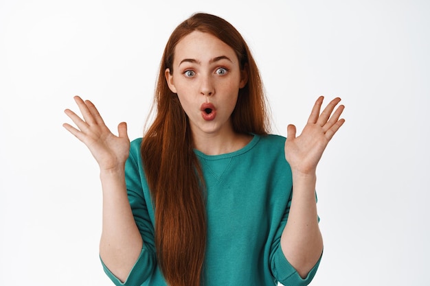 Portrait of surprised caucasian woman with red hair, say wow and stare impressed, spread hands from excitement and amazement, standing over white background.