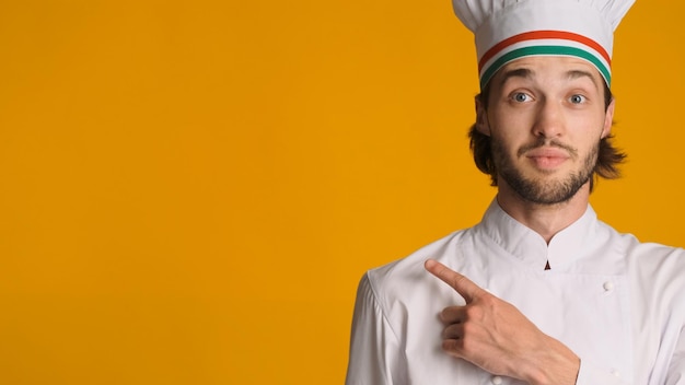 Portrait of surprised chef dressed in uniform pointing away on space for text over colorful background Young man in chef hat showing copy space