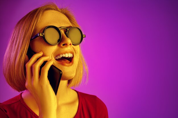 Portrait of surprised confused woman looking at mobile phone isolated over pink neon background