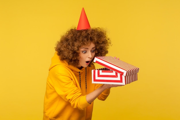 portrait of surprised curious curly-haired woman with party cone hat looking into gift box, opening present and peeking inside with interest, open mouth in amazement. indoor studio shot isolated