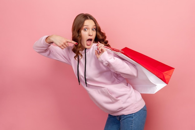 Portrait of surprised curlyhaired teenage girl in hoodie with open mouth pointing finger on paper bags behind back huge discount sale Indoor studio shot isolated on pink background