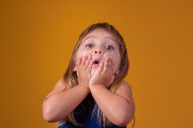 黄色の背景の上に孤立して立っている驚いたかわいい幼児の女の子の子供の肖像画。カメラを見てください。開いた口の近くの手