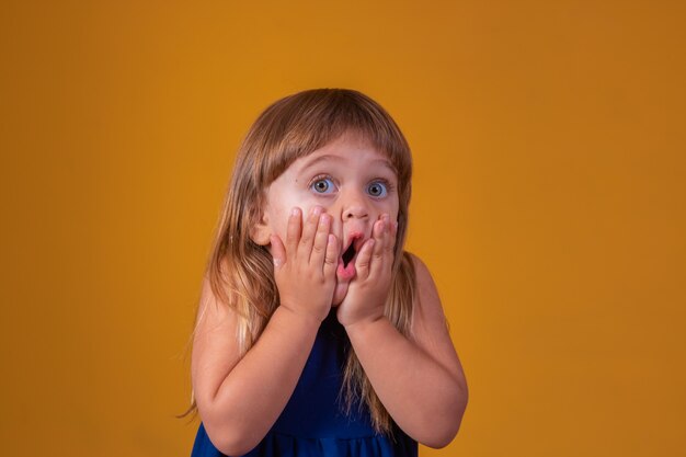 Portrait of surprised cute little toddler girl child standing isolated over yellow background. Looking at camera. hands near open mouth