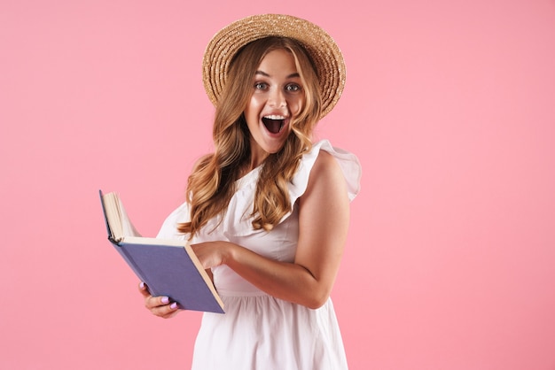 Portrait of a surprised cute young pretty woman posing isolated over pink wall reading book with mouth opened.