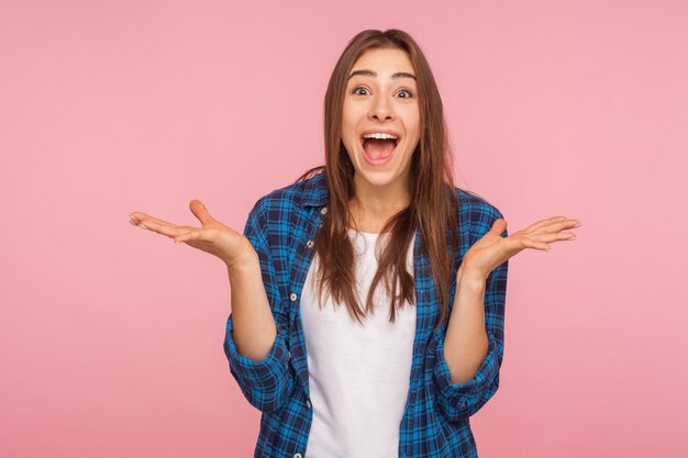 Portrait of surprised emotional funny girl in checkered shirt looking with big eyes, raising hands and screaming in amazement, shocked by sudden event. indoor studio shot isolated on pink background