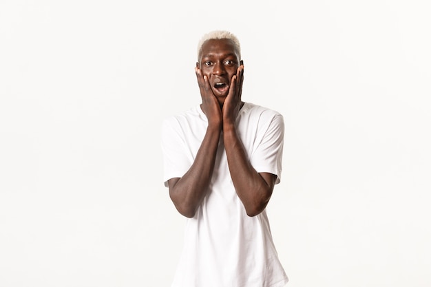 Portrait of surprised and excited african-american blond guy, gasping fascinated, holding hands on face and looking wondered