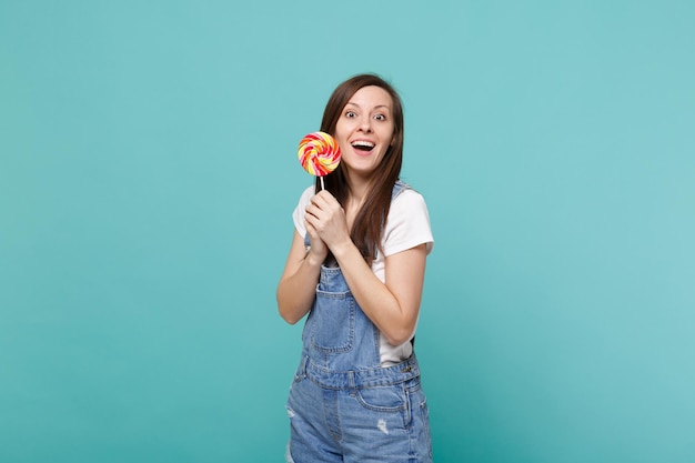 Photo portrait of surprised excited young woman in denim clothes keeping mouth open, holding colorful round lollipop isolated on blue turquoise wall background. people lifestyle concept. mock up copy space.