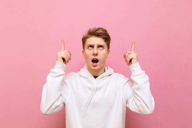 Portrait of surprised guy in white casual clothes stands on a pink background looks up