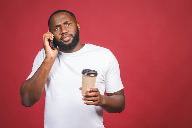 Portrait of surprised handsome African American man with mobile phone and take away coffee cup. Isolated over red background.